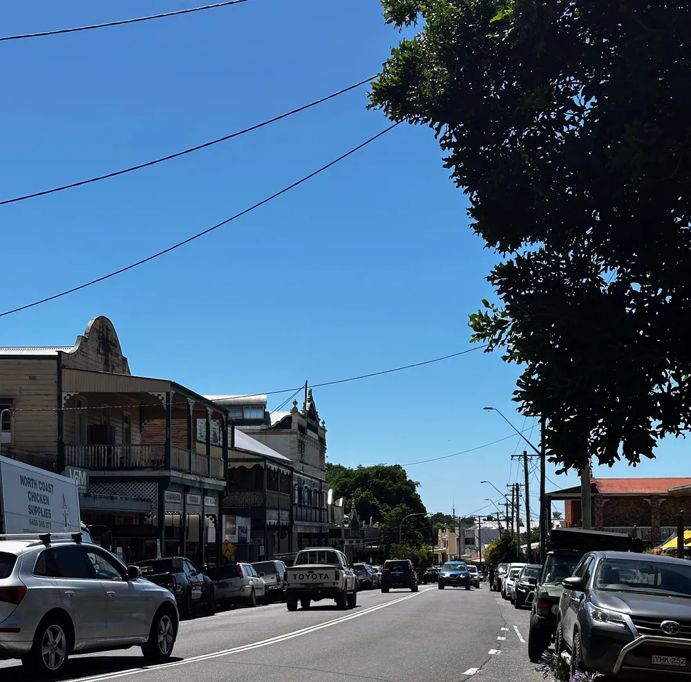 A sunlit small-town main street with iron-lace verandahs and utes parked along the kerb
