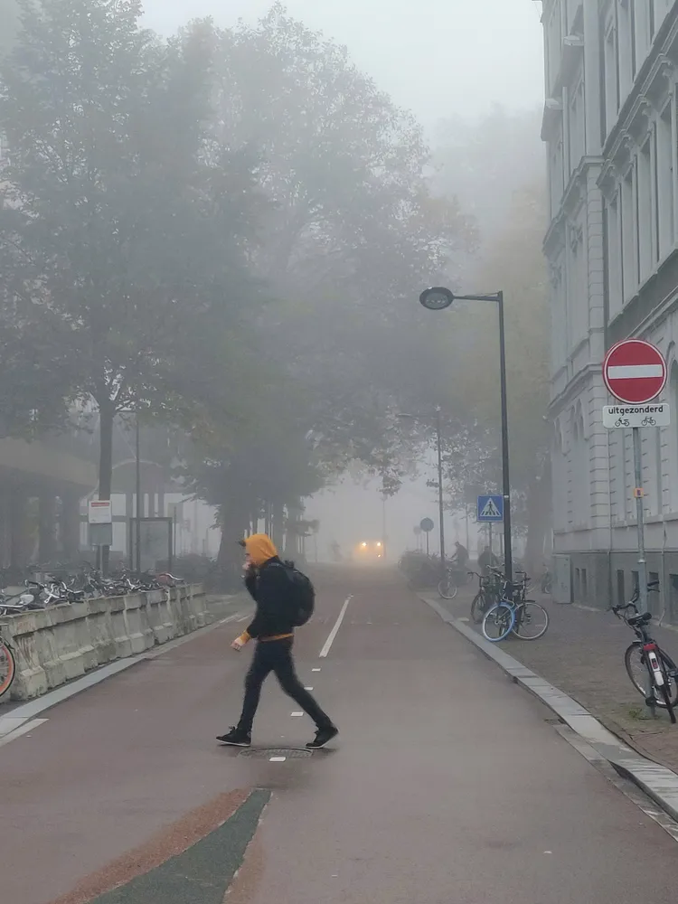 A figure in a yellow hood crossing an empty road in thick fog, a single headlight behind them