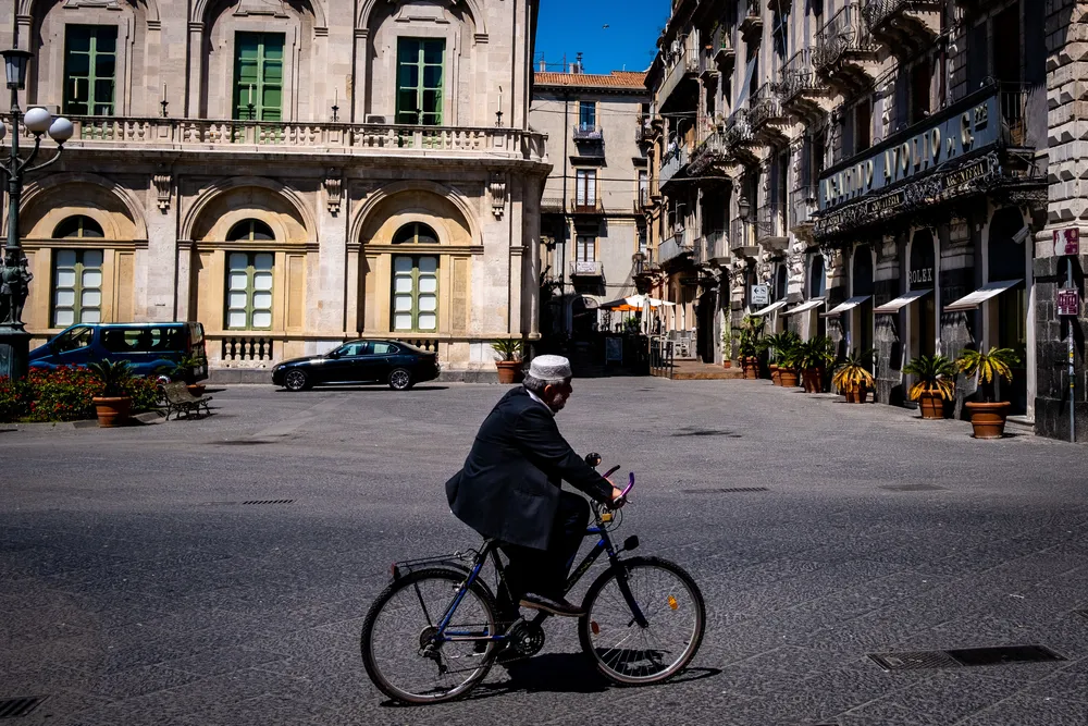 A man in a white cap cycling across an empty sunlit square past shuttered arcades