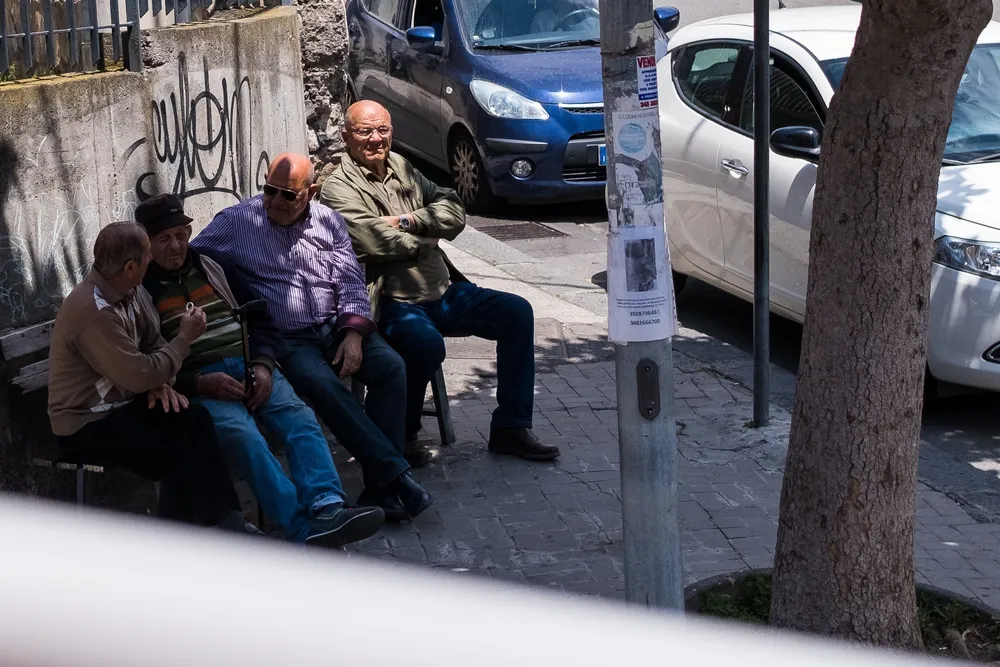 Four older men sitting talking on a bench in the shade, glimpsed between parked cars