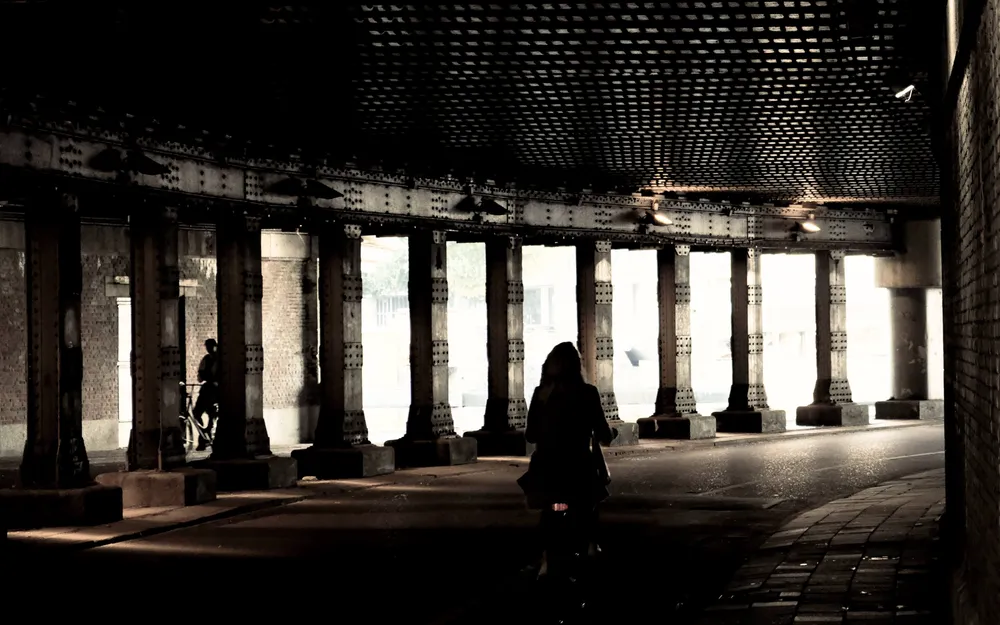 A woman walking through a colonnade of riveted iron pillars under a railway, backlit by daylight