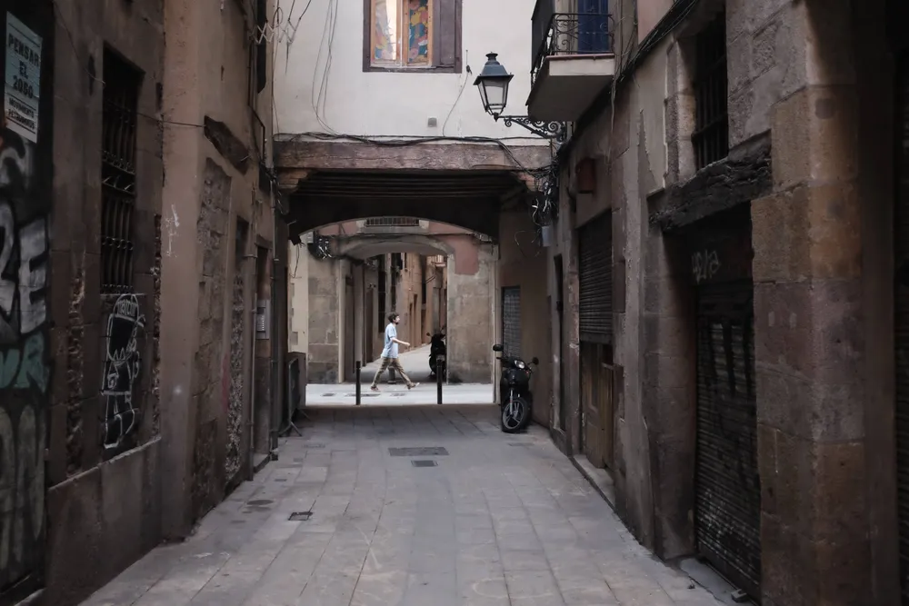 A narrow Barcelona alley in shadow, a single figure crossing the bright gap at the far end