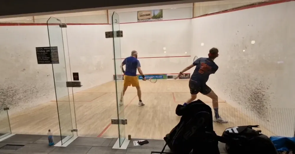 Two players mid-rally, seen through the glass back wall of a court