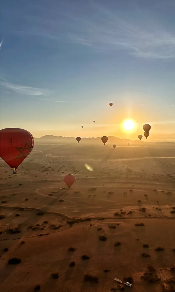 Hot air balloons drifting over desert at sunrise, the nearest one carrying the Moroccan flag